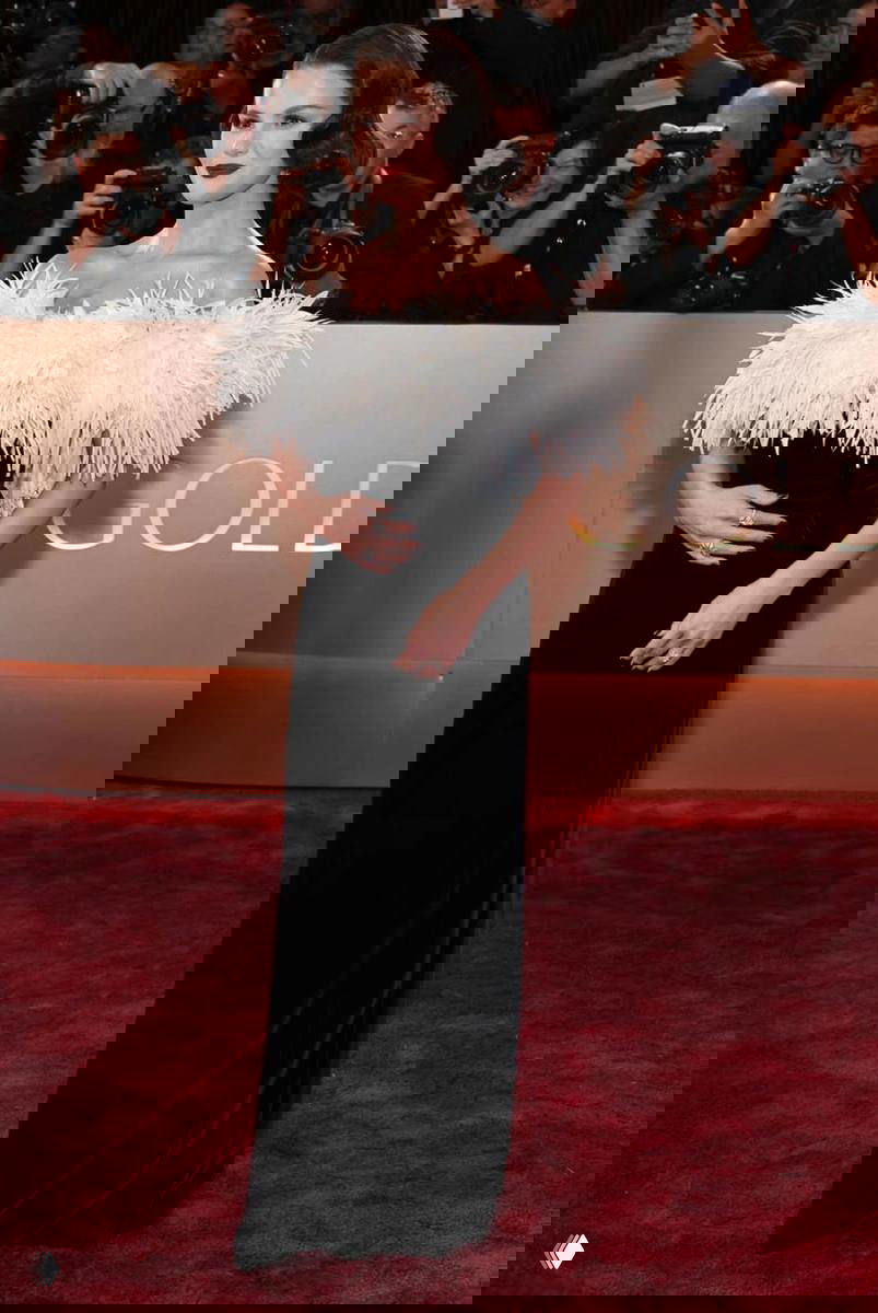 Полноразмерный кадр на красной дорожке: женщина в чёрном платье с белыми перьями на лифе, рука у живота; на заднем плане фотографы и гости.