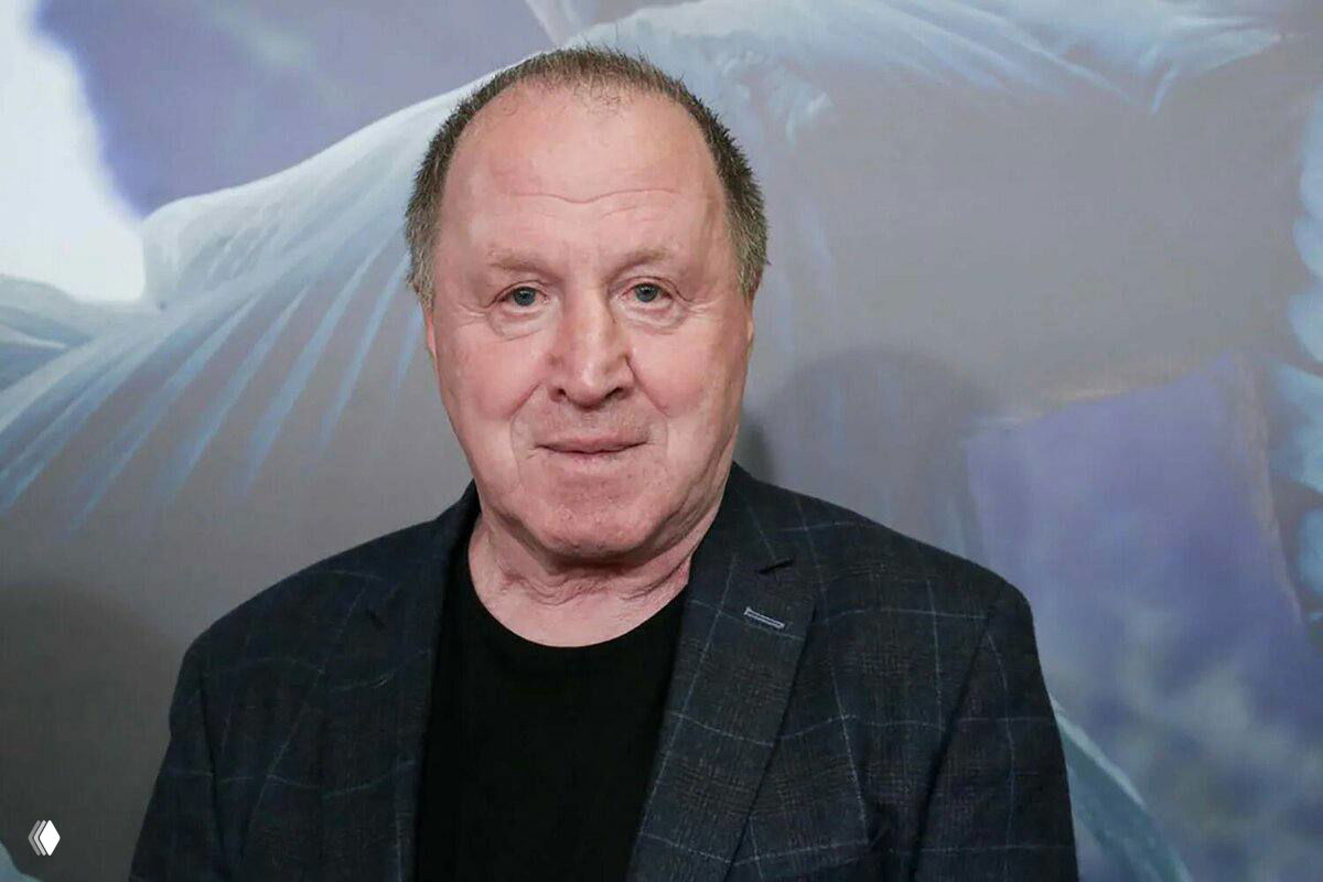 Портрет пожилого мужчины в пиджаке на нейтральном фоне, кадр с выражением спокойного лица в официальной обстановке.