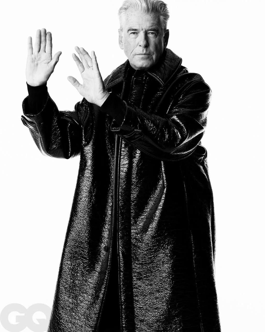 Полнофигурный чёрно‑белый снимок: мужчина в глянцевом плаще поднимает руки в драматичной позе, подчёркнутая модная эстетика.
