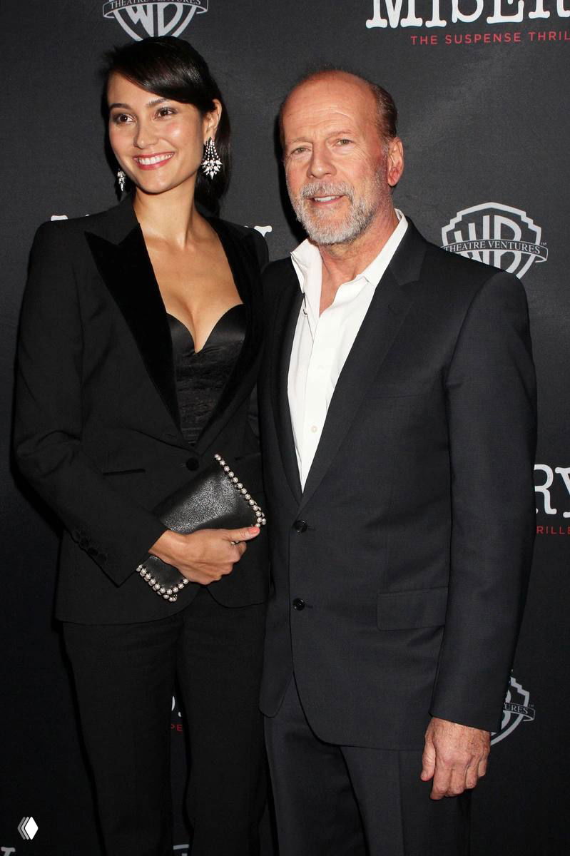 Пара на красной дорожке: мужчина и женщина в вечерней одежде стоят рядом и позируют на фоне баннера события с логотипами.