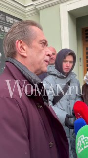 Крупный план мужчины у входа в МХТ среди людей и микрофонов СМИ; на переднем плане разноцветные микрофоны и водяной знак WOMAN.ru