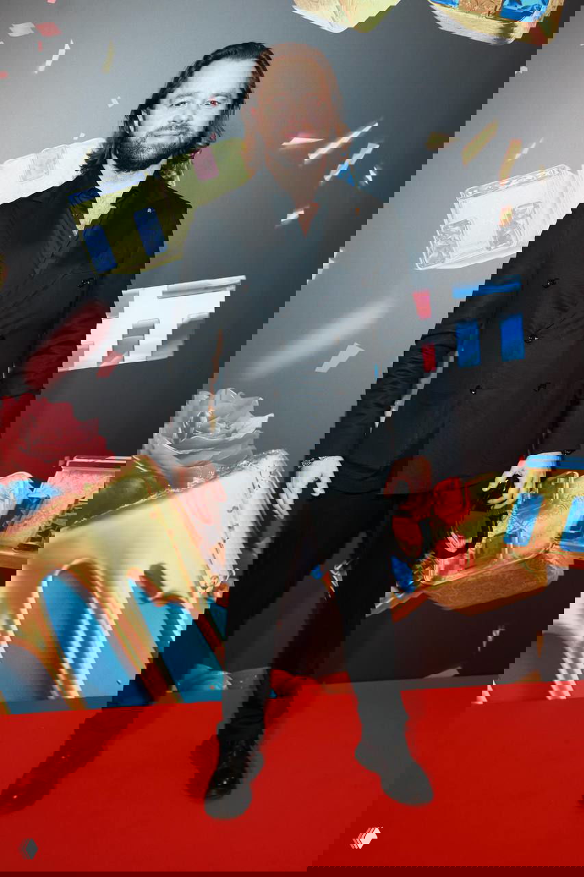 Виталий Гогунский в тёмном костюме стоит на красной дорожке перед яркой декорацией с конфетами и логотипом, полный рост, официальное фото.