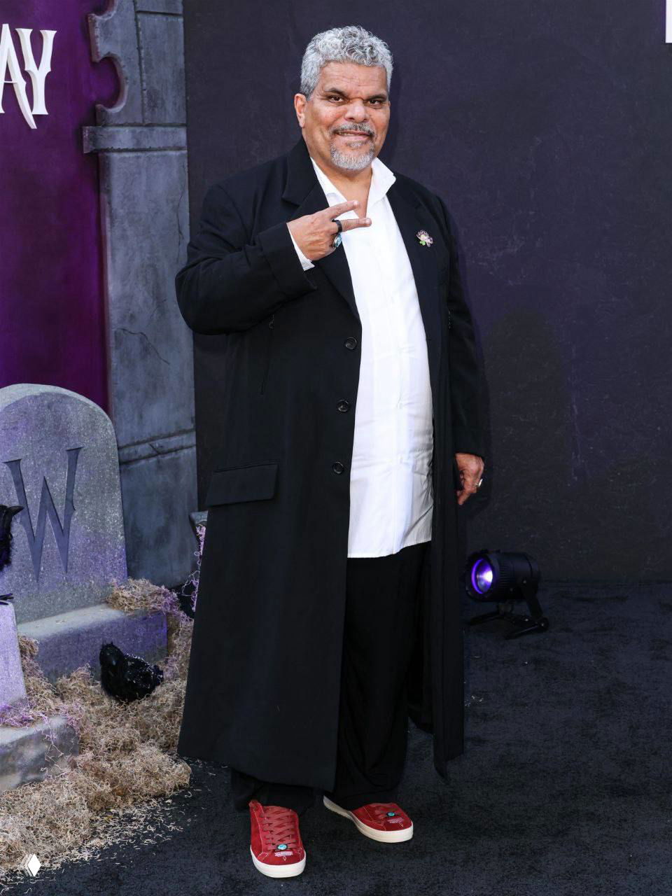 Луис Гусман в длинном черном пальто и красных кедах позирует на фоне фотозоны «Уэнсдей» на мероприятии в Лос‑Анджелесе.