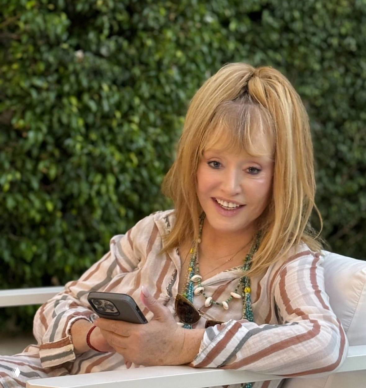 Средний план: женщина со светлыми волосами в полосатой блузке улыбается и держит смартфон на фоне зелёной живой изгороди, интерьер на улице