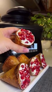 Крупный план руки с рулетиком, наполненным кремом и зернами граната; фон — плитка и праздничная сервировка на столе.