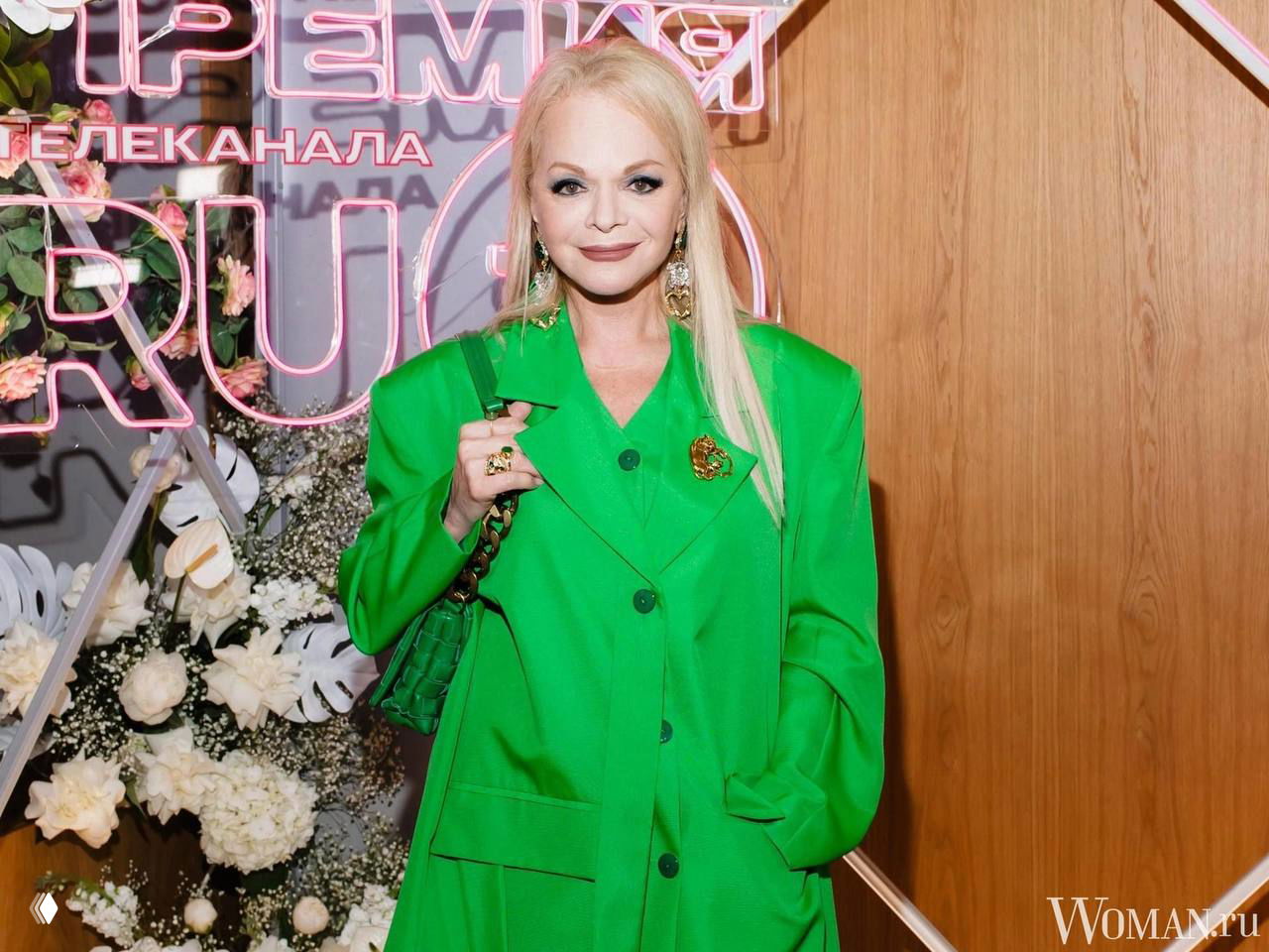 Женщина в ярко‑зелёном наряде на светском мероприятии, позирует на фоне цветочной декорации и неоновых букв, официальный кадр