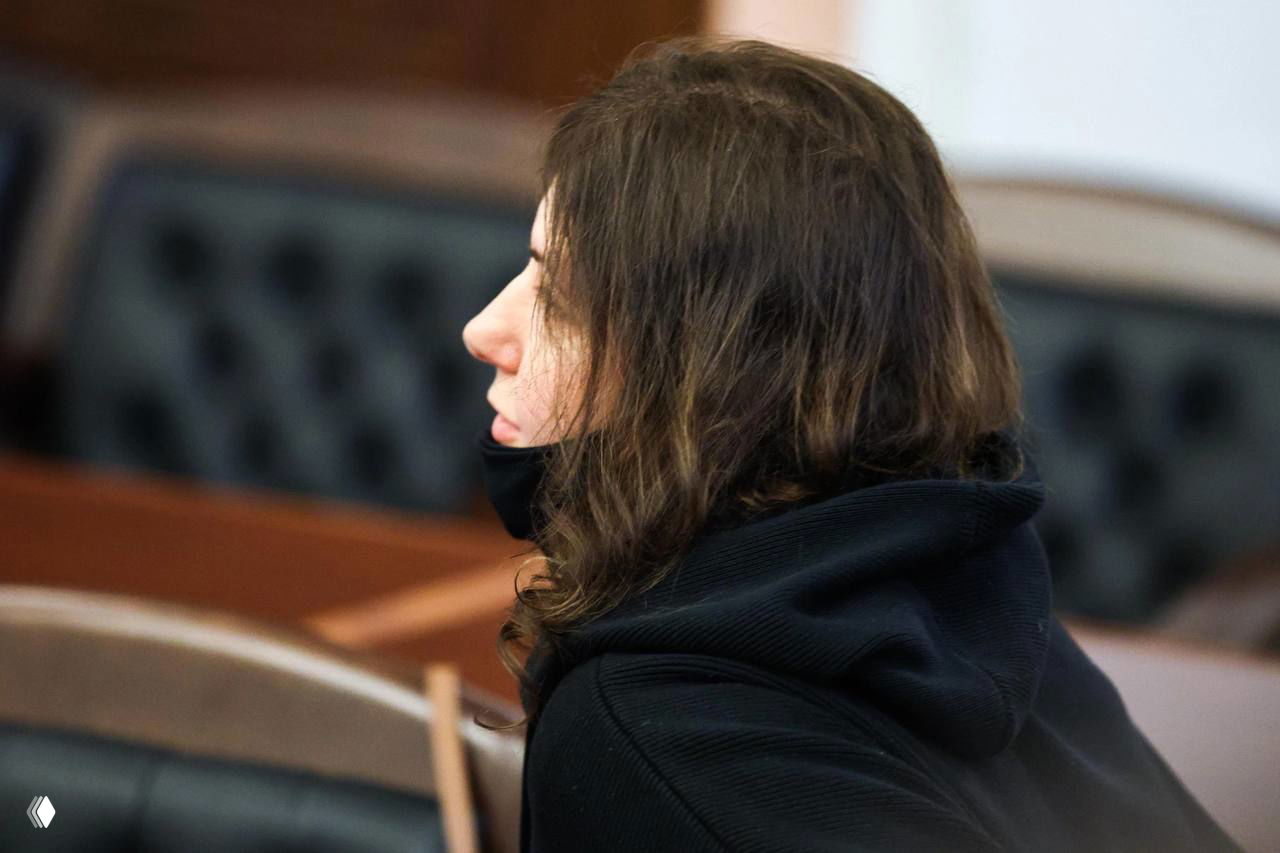 Профиль женщины в тёмной одежде и с маской под подбородком в зале заседаний суда во время слушания по делу