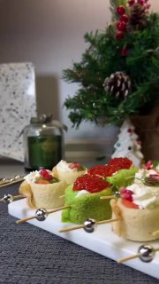 Несколько канапе и рулетиков разной формы на белой тарелке рядом с веткой ели и праздничными украшениями на фоне.