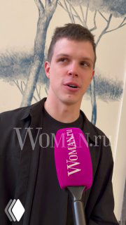 Мужчина в тёмном пиджаке даёт интервью у микрофона с логотипом Woman.ru на фоне художественной настенной росписи.