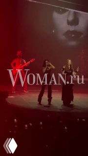 Снимок с концерта «Тату» в Москве: два участника на сцене, гитара и большой экран с чёрно‑белым портретом, публика и логотип Woman.ru в кадре