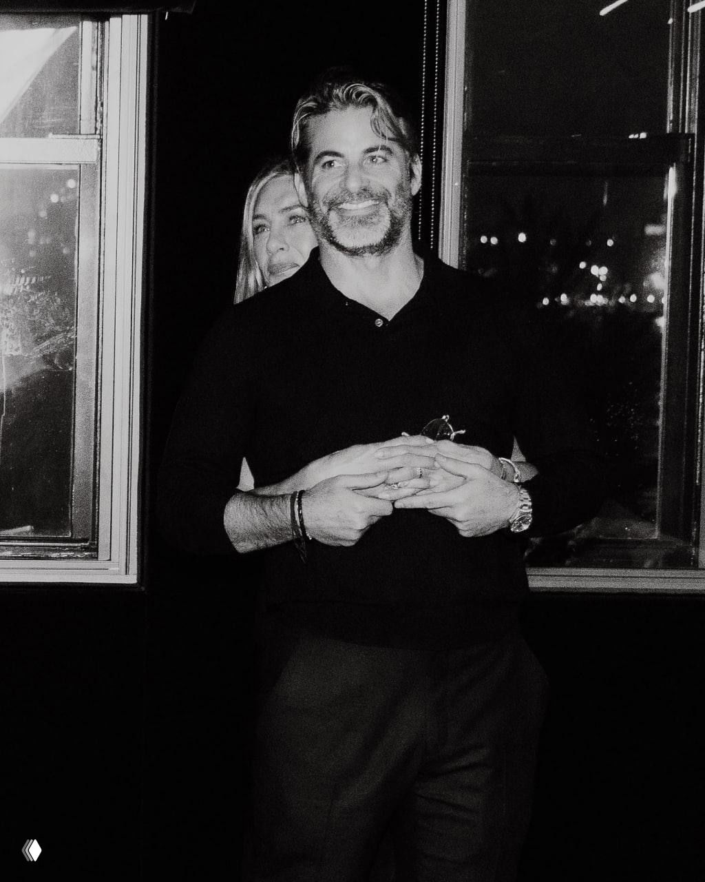 Чёрно‑белое фото: улыбающийся мужчина в свитере стоит на переднем плане, женщина (Дженнифер Энистон) позади обнимает его за талию — совместный кадр в романтической обстановке.