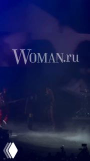 Фото с концерта «Тату»: сцена в дымке, группа на фоне синего освещения и силуэты музыкантов, логотип Woman.ru виден поверх изображения