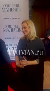 Женщина в тёмной одежде на красной дорожке у постера фильма «Огненный мальчик», кадр с премьеры с логотипом Woman.ru и фотозоной.