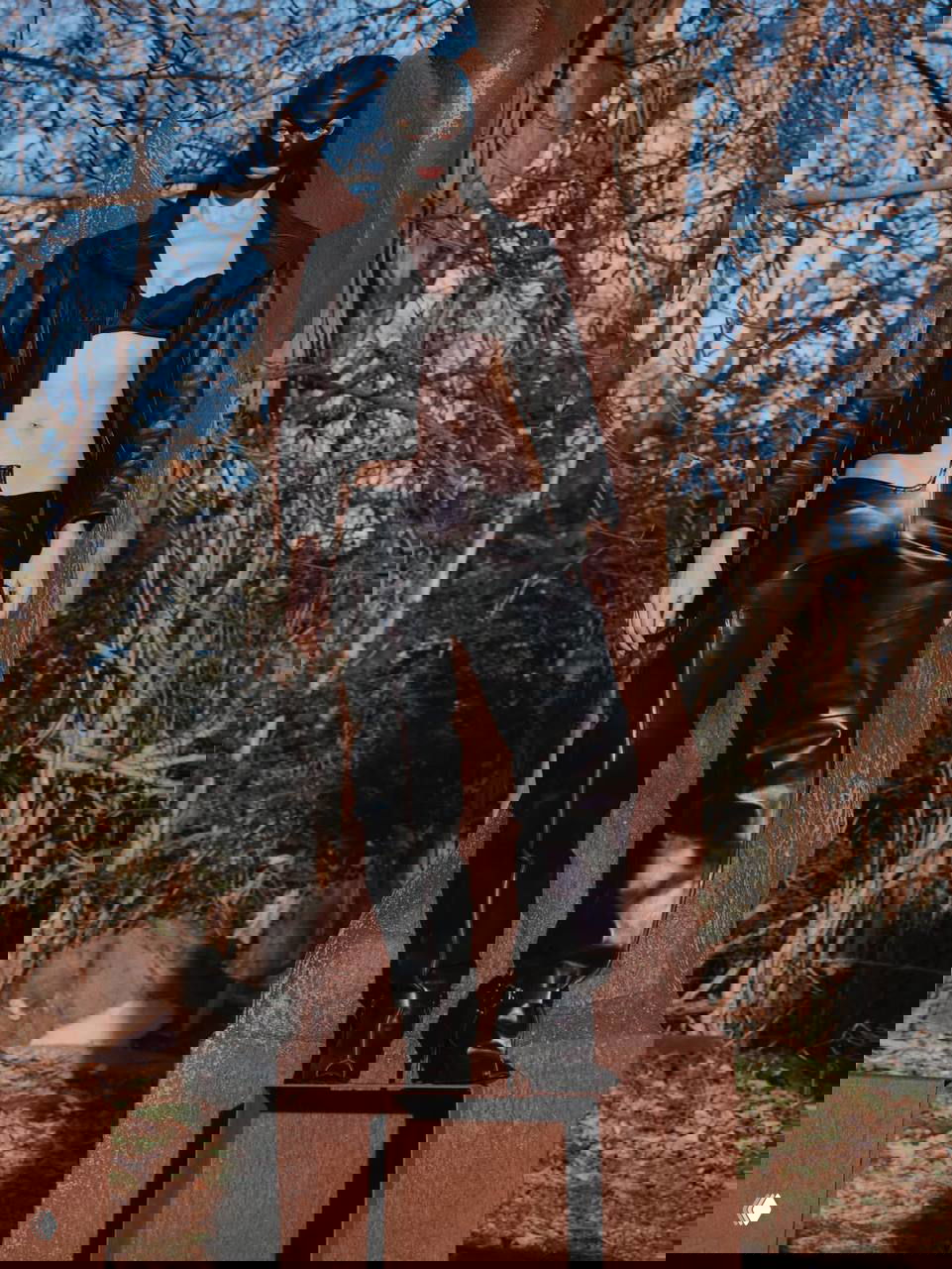 Фигура модели в чёрных кожаных штанах и маске на фоне деревьев — контрастная уличная постановка с элементами авангарда и дерзкого стиля.