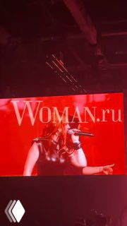 Фото с выступления «Тату»: крупный план певицы на большом экране за сценой, яркая красная подсветка и узнаваемый водяной знак Woman.ru