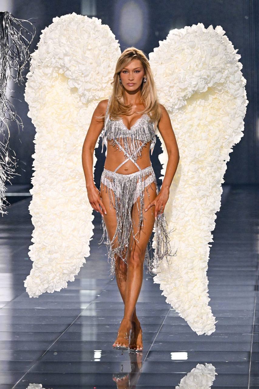 Модель на подиуме в наряде в стиле Victoria’s Secret с большими белыми крыльями, блестящий комплект и отражение на глянцевом полу показа