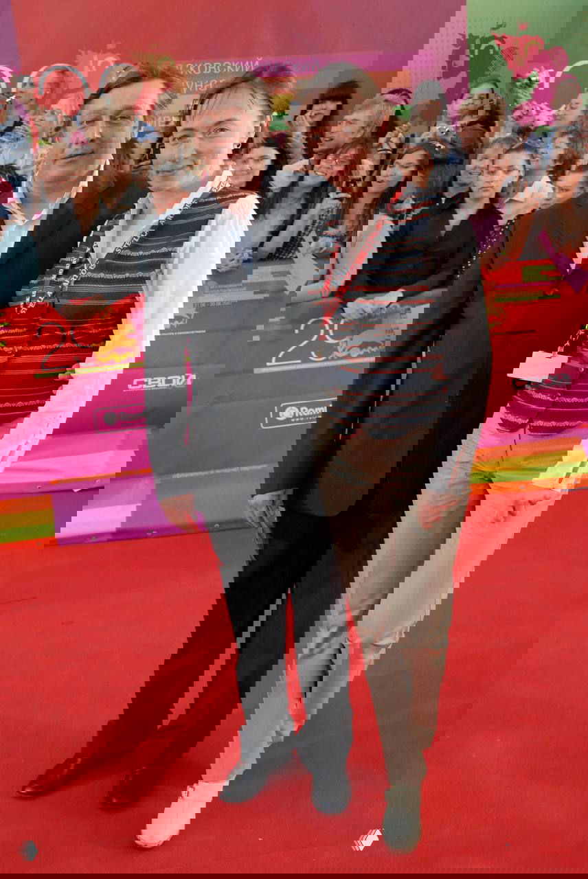 Мужчина и женщина позируют на красной дорожке перед публикой и яркими баннерами — кадр со светского мероприятия.