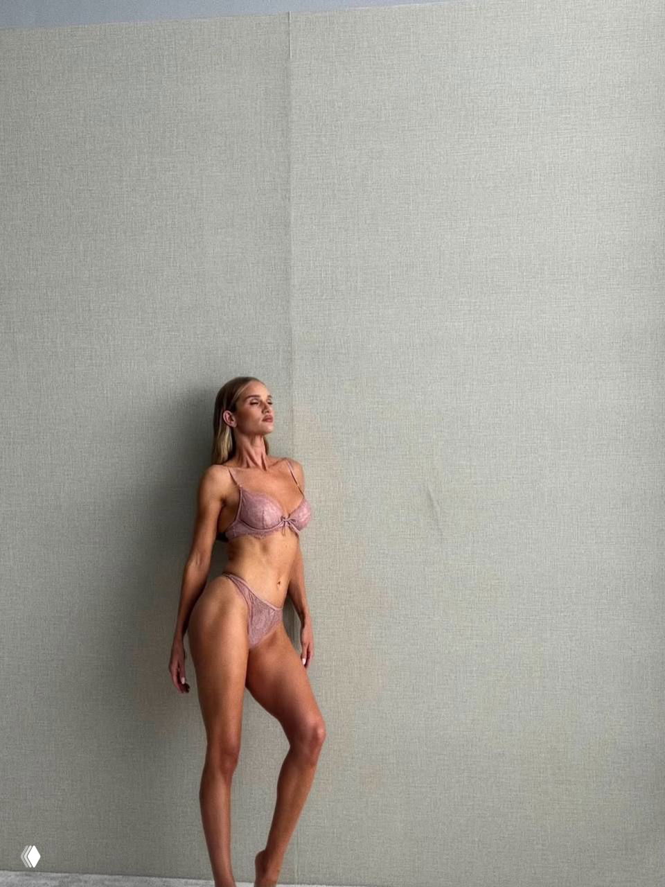 Ростовой кадр Роузи Хантингтон-Уайтли в розовом белье на однотонном фоне; модель стоит, демонстрируя силуэт и фактуру комплекта в студийном освещении.