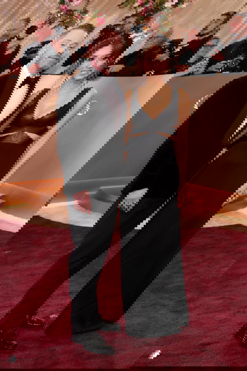 Пара позирует вместе на красной дорожке у декоративной фотозоны церемонии; вокруг — фотографы и гости.