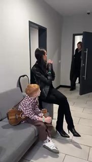 Мальчик с гитарой сидит на диване в коридоре студии, рядом женщина в чёрном с высокими каблуками, в дверном проёме взрослый.