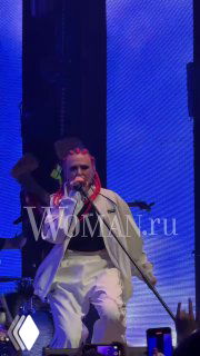 Крупный план солиста группы «Вирус!» на сцене VK Gipsy: микрофон и эмоциональное исполнение, белая одежда и LED‑подсветка, заметен логотип Woman.ru