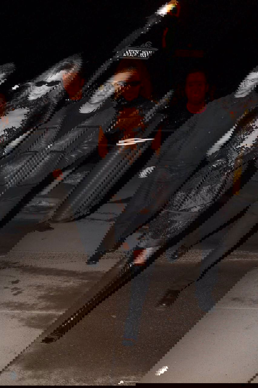 Модель в длинных черных лаковых сапогах и полупрозрачном кружевном платье, идет по вечерней улице зимнего курорта в ночном освещении.