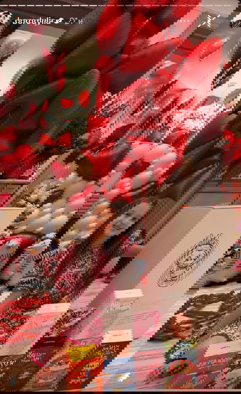 Девочка в нарядном платье сидит на полу среди подарков, брендовой упаковки и надувных игрушек‑пингвинов на фоне сердечных шаров.