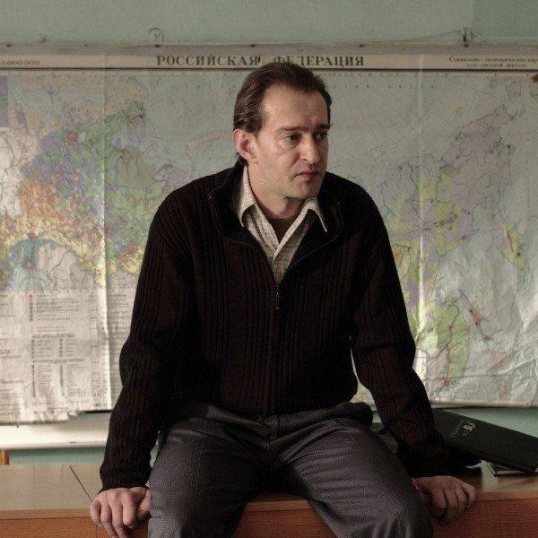 Мужчина в тёмном пуловере на фоне большой карты, поза сидя, образ делового или служебного характера, интерьер в офисном стиле.