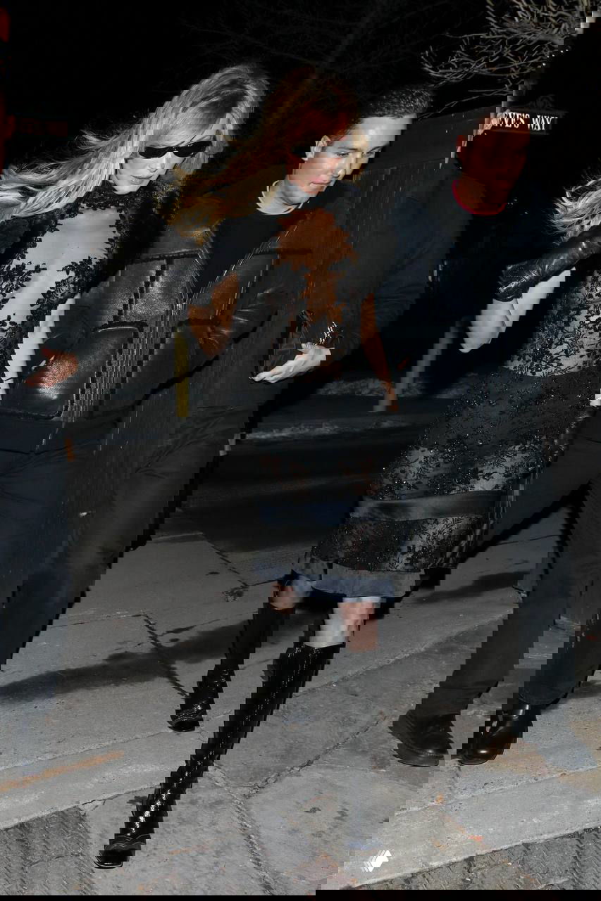 Модель в черном кружевном платье и солнцезащитных очках шагает по ночной улице курорта рядом с сопровождающими.