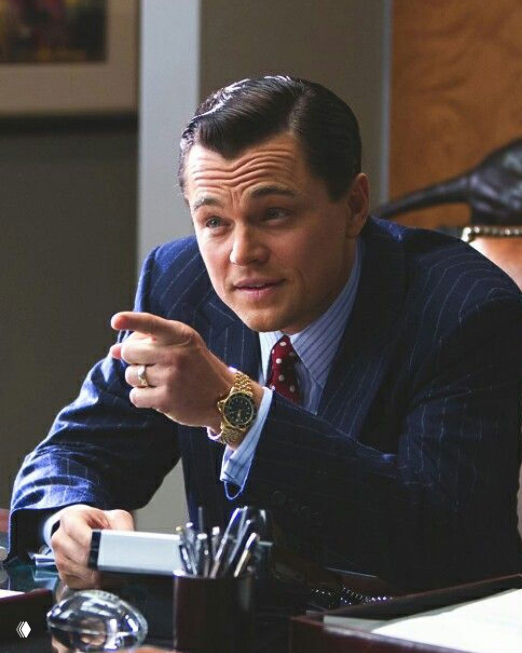 Кадр с ДиКаприо в деловом костюме, сцена из фильма с жестикуляцией персонажа за рабочим столом, выражение лица сосредоточенное и уверенное.