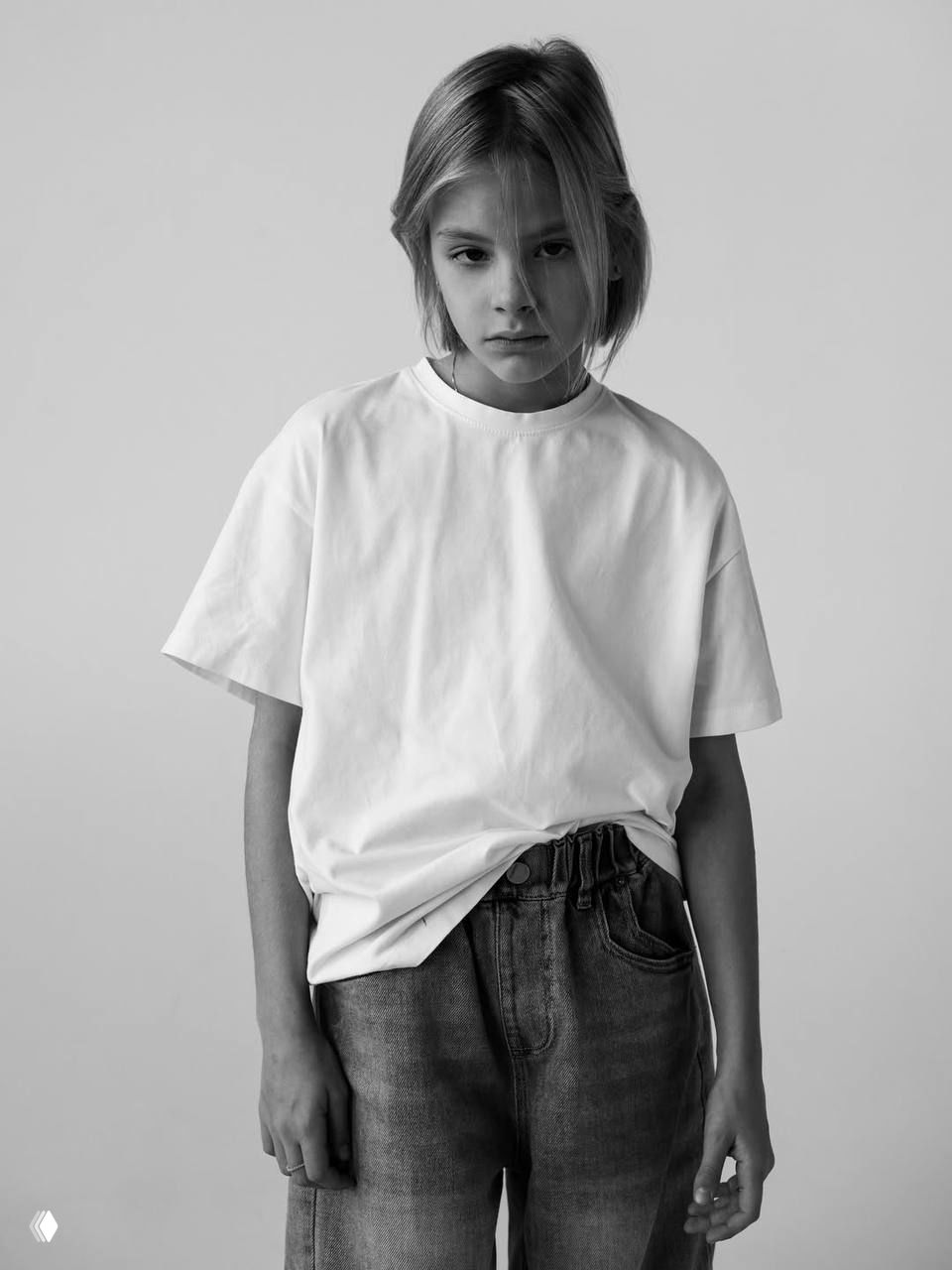 Черно‑белая фотография девочки в три‑четверти, руки у лица, искренняя улыбка и лёгкая игра эмоциями в студии.