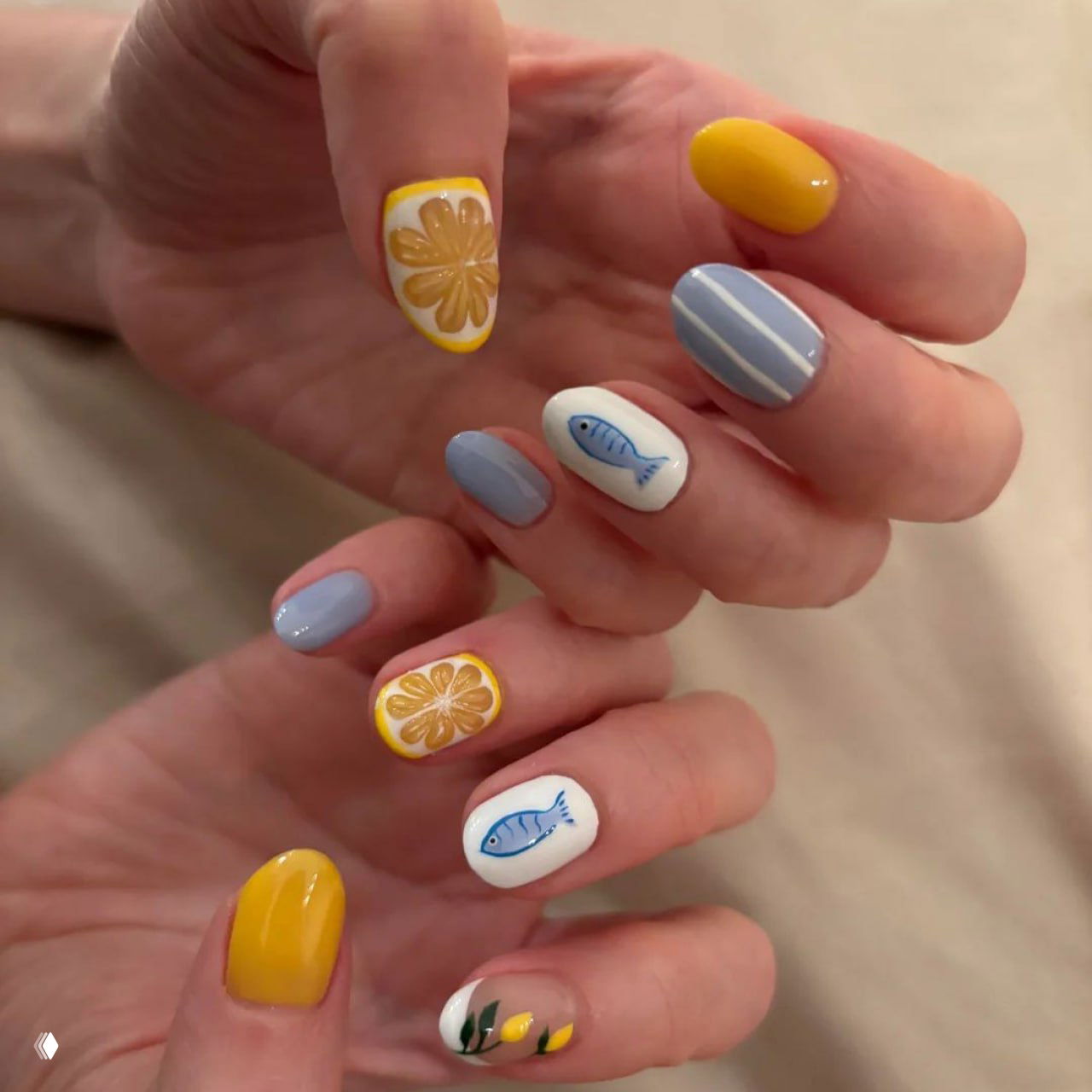Крупный план пальцев с маникюром: сочетание нежно-голубых, белых и жёлтых оттенков, рисунки рыбы и дольки цитруса на ногтях.