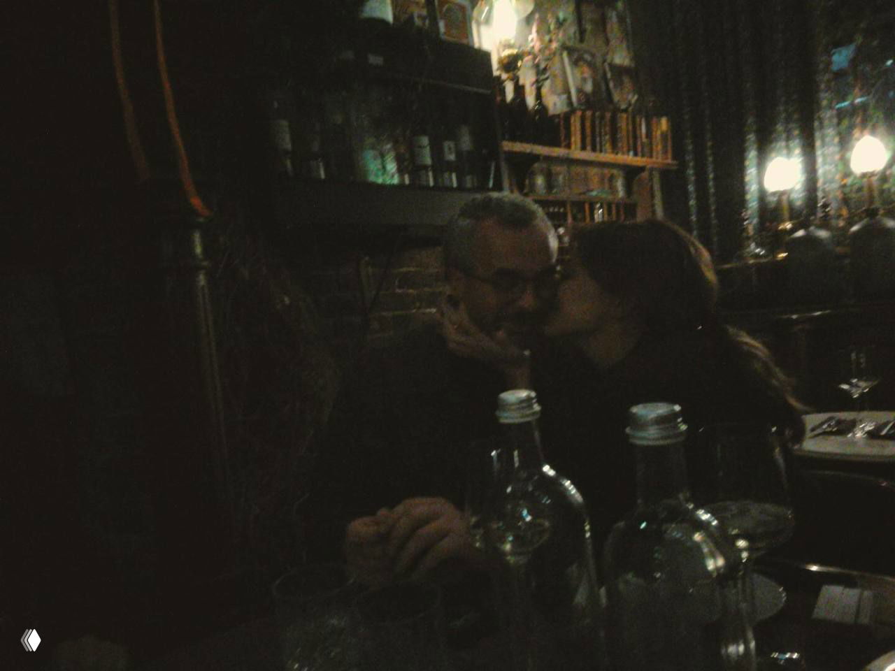 Тёмный атмосферный снимок в баре: пара целуется за столом, на переднем плане бутылки и бокалы, приглушённое освещение и интерьер заведения.
