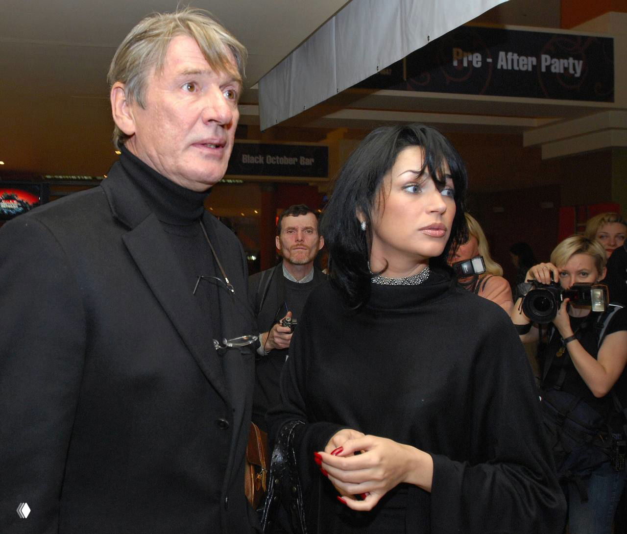 Юлия Абдулова и мужчина в тёмной одежде на светском мероприятии, фотографы и зрители видны на заднем плане в зале, кадр сделан в помещении.