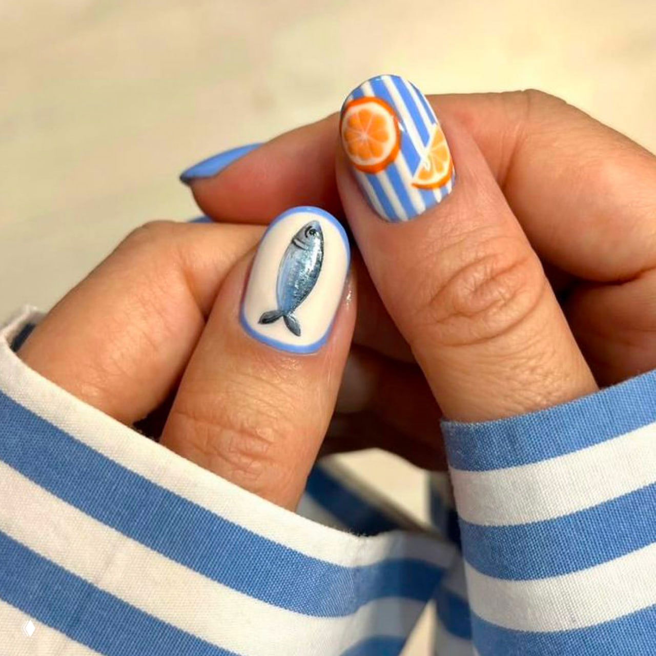 Близкий план ногтей с синими и белыми полосами, маленькой рыбкой и апельсиновым рисунком — морской летний стиль маникюра.