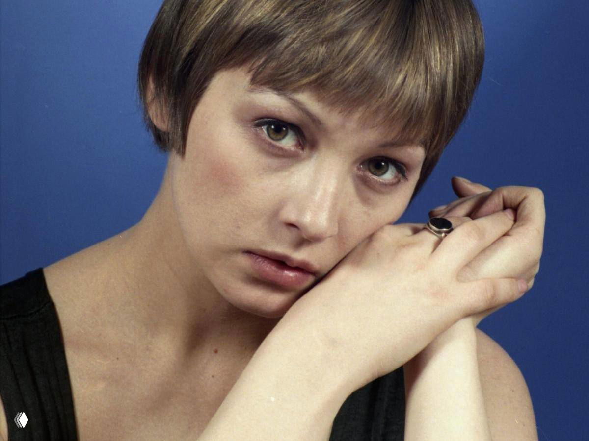 Крупный план портрета женщины с короткой стрижкой на синем фоне, задумчивый взгляд и сложенные у лица руки.