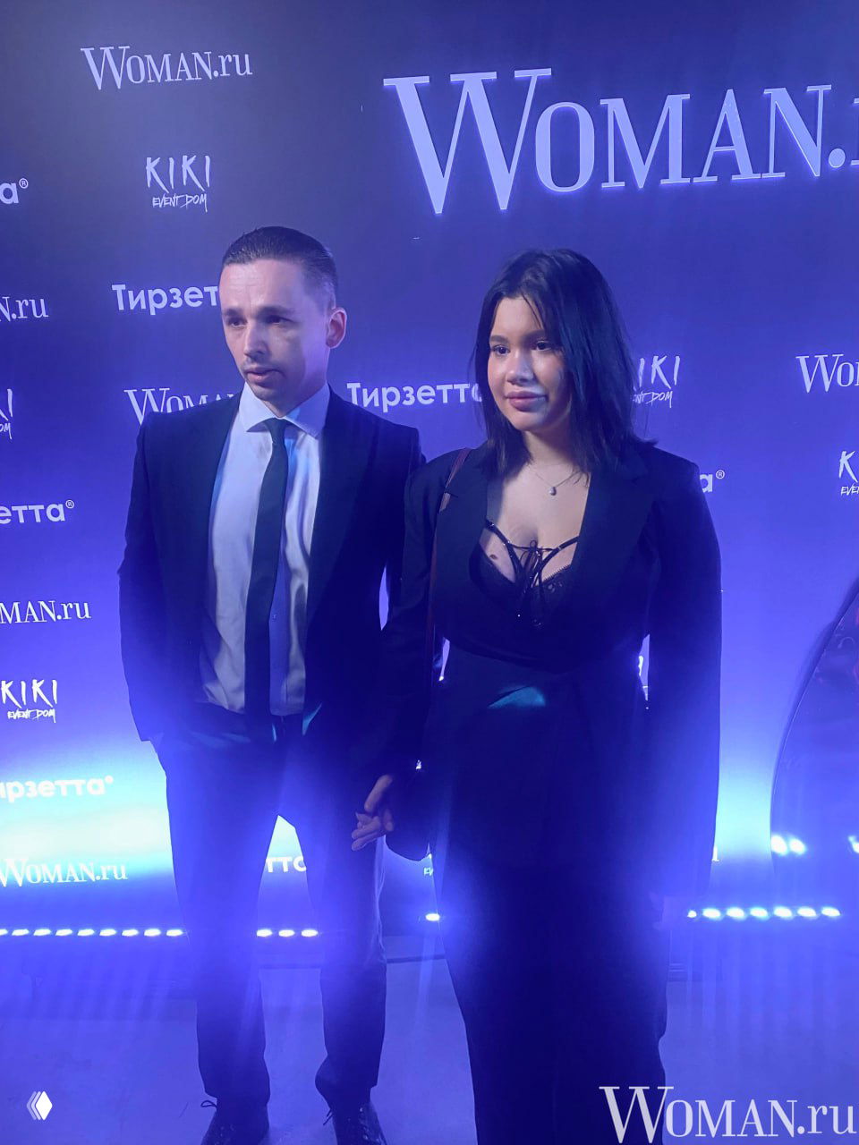 Крупный план: Полина и Артур стоят у бренд-стены Woman.ru на вечеринке Celebrity Party, видны вечерние костюмы и синяя подсветка.