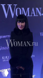 Женщина в тёмной верхней одежде стоит у пресс-волла с логотипом Woman.ru во время праздничного мероприятия издания Woman.ru.