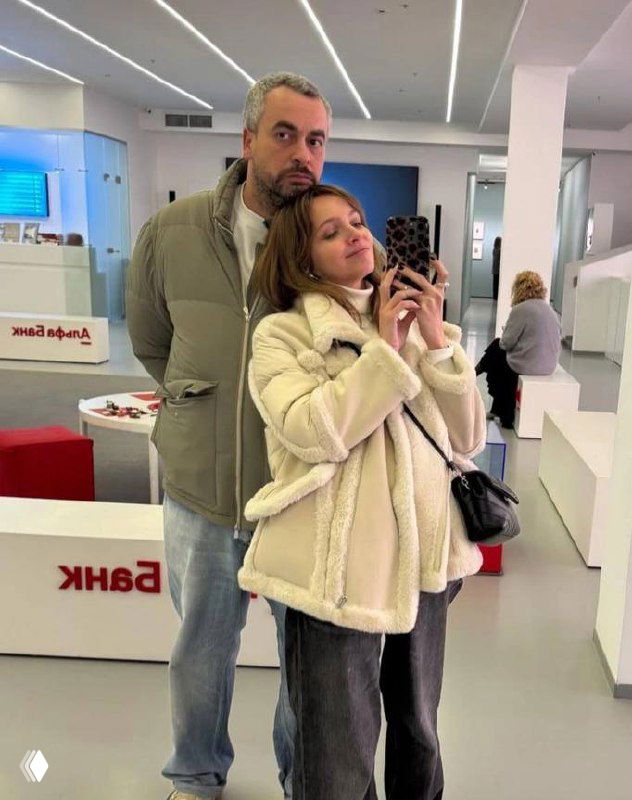 Селфи в галерейном интерьере: женщина в светлой дублёнке делает фото на телефон, рядом высокий мужчина, выставочные объекты и минималистичный интерьер.