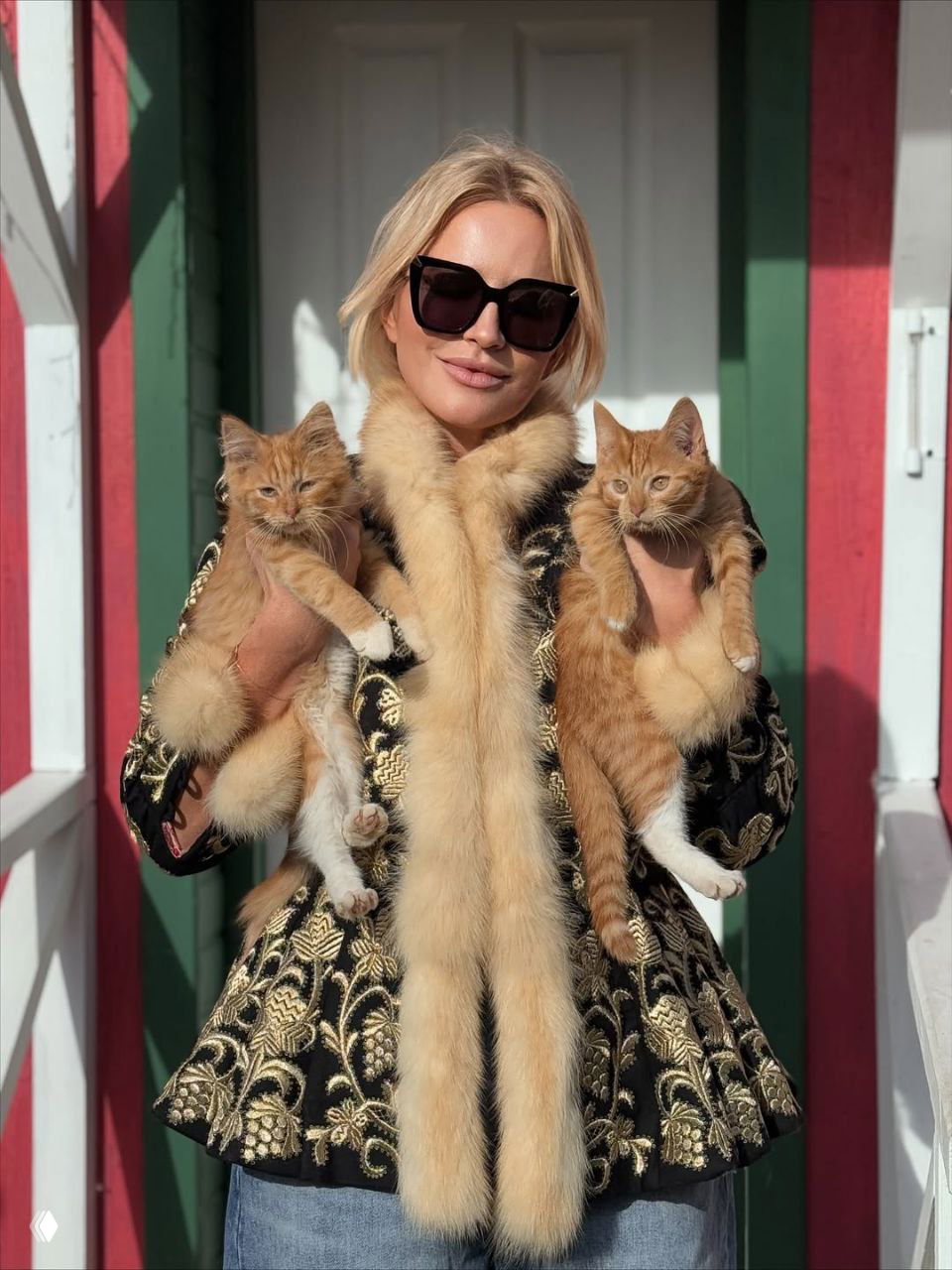 Женщина в роскошном жакете с меховым воротником и солнцезащитных очках держит двух рыжих котят на фоне деревянного дома.