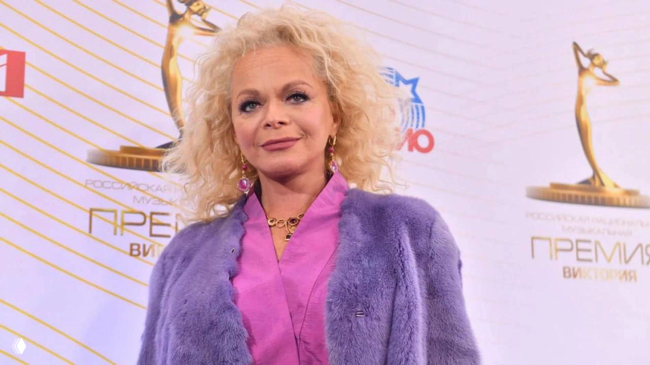 Лариса Долина на красной дорожке в ярком образе: портрет певицы в фиолетовой шубе на фоне логотипа премии, крупный план в светской обстановке.