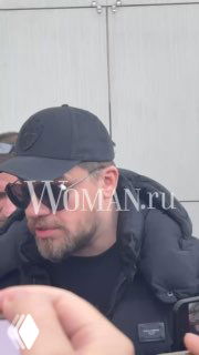 Мужчина в тёмной кепке и тёмных очках во время съёмки/интервью; на фото видна водяная метка Woman.ru и часть оборудования съёмочной группы.