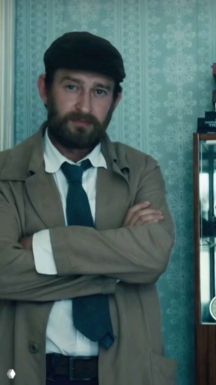 Вертикальный кадр мужчины в плаще и галстуке, руки скрещены, интерьер жилого пространства с винтажными обоями, атмосферное освещение.