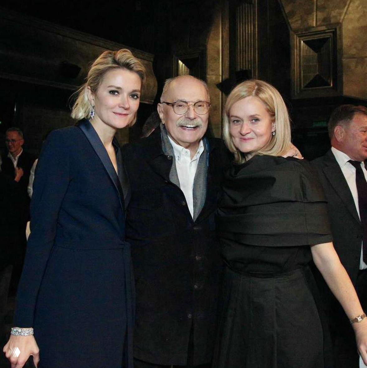 Фотография с тремя людьми в тёплой обстановке — гости улыбаются, вокруг столы и элементы ресторанной сервировки.