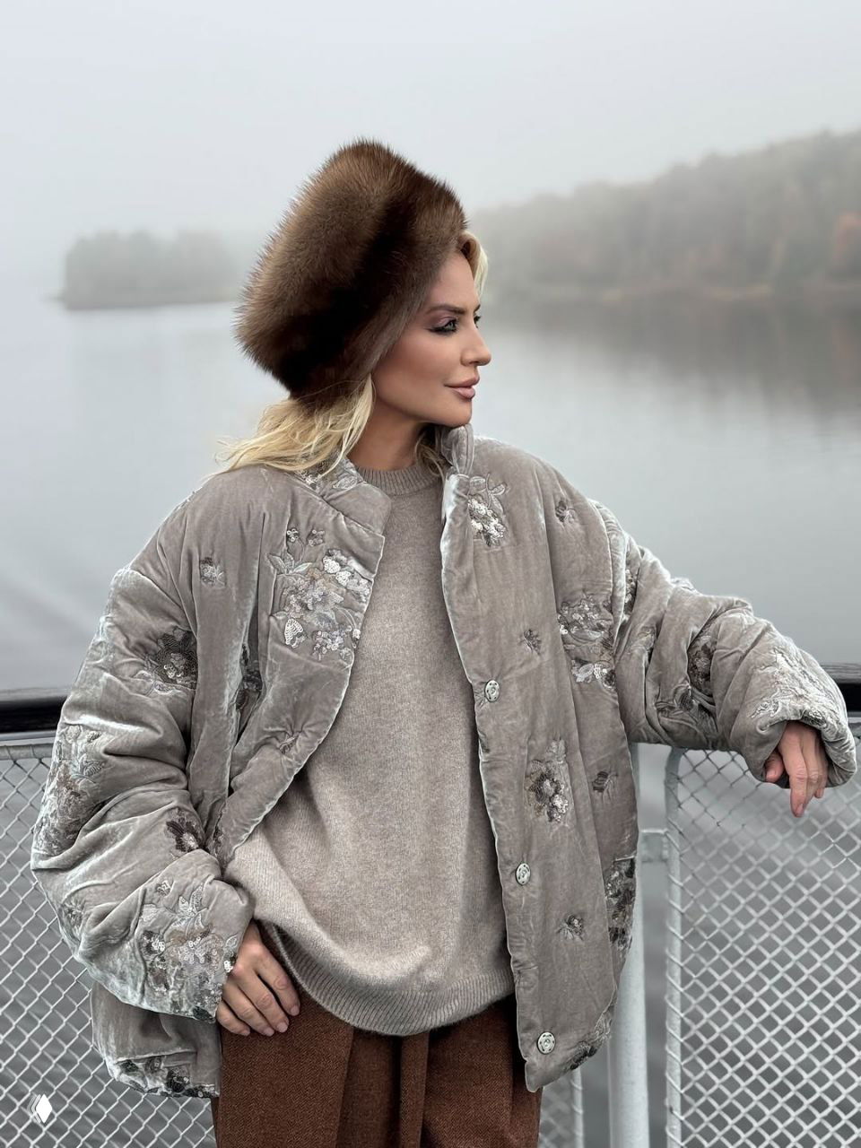 Женщина в объёмной серой стёганой куртке с вышивкой и меховой шапке стоит у туманного озера, спокойный водный фон и осенние берега.