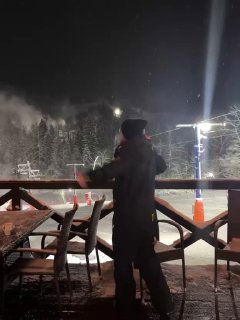 Ночной кадр на открытой террасе возле освещённой лыжной зоны: силуэт человека в тёплой одежде, вокруг заметна зимняя ночь и лёгкая подсветка трассы.