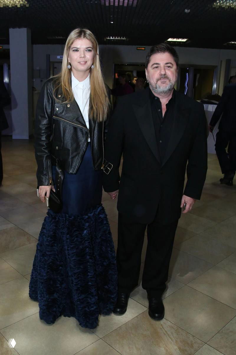 Мужчина и женщина в вечерней одежде позируют в фойе светского мероприятия; пара стоит рядом, держатся за руки, виден интерьер зала