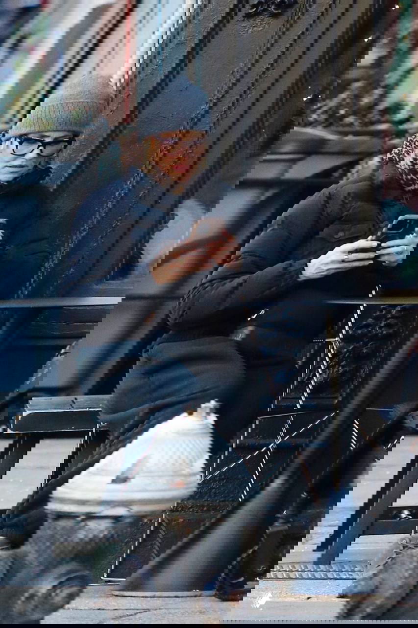 Кэмерон Диаз сидит на уличной скамейке в тёплой одежде и шапке, держит телефон; фото папарацци в городском окружении во время прогулки.