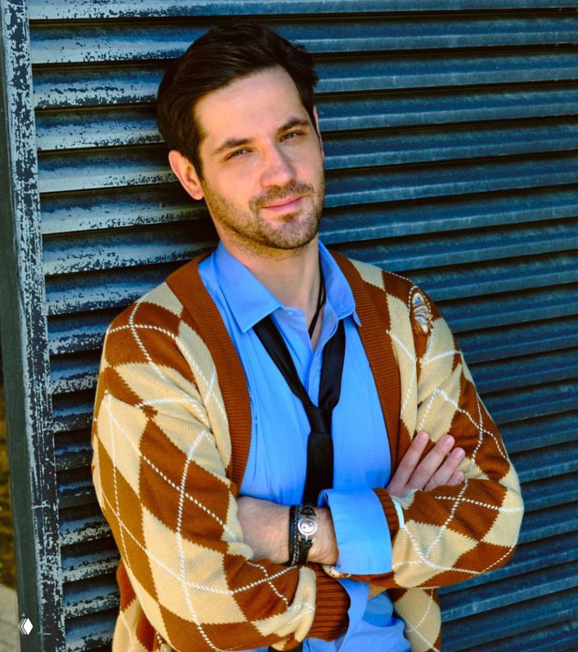 Портрет мужчины в кардигане с ромбическим рисунком и голубой рубашке; полуближний план на фоне металлической жалюзи, непринуждённая поза.