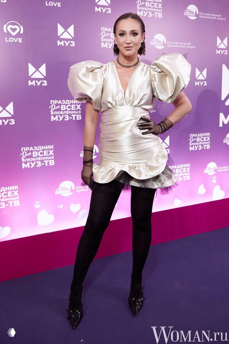 Фотография с фиолетовой фотозоны: женщина в коротком сатиновом платье с объёмными рукавами и тёмными колготками, необычный модный силуэт.